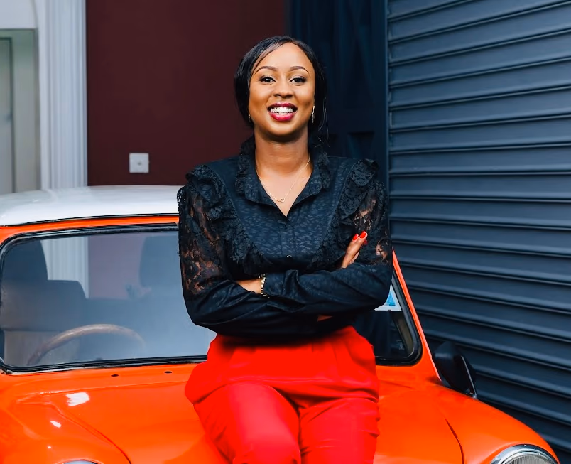 Joy Mido seated in a black blouse and red trousers
